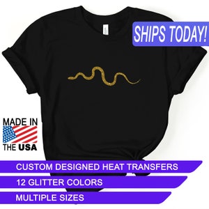 May include: Black t-shirt with a gold glitter snake design. The shirt says "SHIPS TODAY!" in a blue box. It also says "MADE IN THE USA" with an American flag. The text "CUSTOM DESIGNED HEAT TRANSFERS", "12 GLITTER COLORS", and "MULTIPLE SIZES" are in blue boxes.