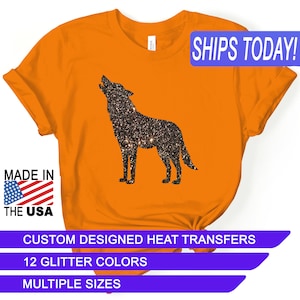 May include: Orange t-shirt with a black glitter wolf silhouette design. The text "SHIPS TODAY!" is in a blue rectangle at the top right corner. The text "MADE IN THE USA" is in a red, white, and blue rectangle at the bottom left corner. The text "CUSTOM DESIGNED HEAT TRANSFERS" is in a purple rectangle at the bottom center. The text "12 GLITTER COLORS" is in a purple rectangle below the previous text. The text "MULTIPLE SIZES" is in a purple rectangle below the previous text.