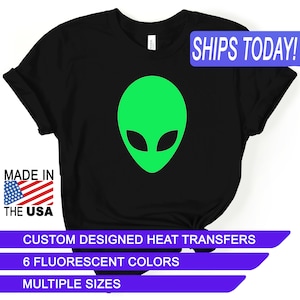 May include: Black t-shirt with a green alien head graphic. The text "SHIPS TODAY!" is in a blue box at the top of the image. The text "MADE IN THE USA" is in a red, white, and blue box at the bottom left of the image. The text "CUSTOM DESIGNED HEAT TRANSFERS", "6 FLUORESCENT COLORS", and "MULTIPLE SIZES" are in purple boxes at the bottom of the image.