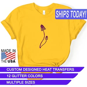 May include: Yellow t-shirt with a purple glitter mushroom design. The shirt says "SHIPS TODAY!" in blue text. The shirt also says "MADE IN THE USA" with a small American flag. The shirt also says "CUSTOM DESIGNED HEAT TRANSFERS", "12 GLITTER COLORS", and "MULTIPLE SIZES" in purple text.