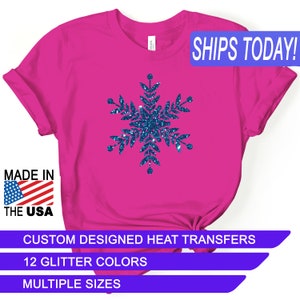 May include: A pink t-shirt with a blue glitter snowflake design. The shirt says "SHIPS TODAY!" in blue text. The shirt also says "MADE IN THE USA" and "CUSTOM DESIGNED HEAT TRANSFERS". It also says "12 GLITTER COLORS" and "MULTIPLE SIZES".