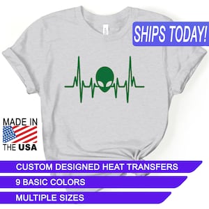 May include: A gray t-shirt with a green alien head inside a heartbeat graphic. The text "SHIPS TODAY!" is in a blue box at the top of the image. The text "MADE IN THE USA" is in a red, white, and blue box at the bottom left of the image. The text "CUSTOM DESIGNED HEAT TRANSFERS" is in a purple box at the bottom of the image. The text "9 BASIC COLORS" is in a purple box below the previous text. The text "MULTIPLE SIZES" is in a purple box below the previous text.