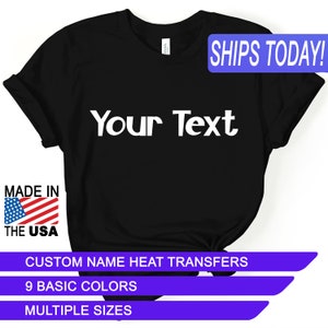 May include: Black t-shirt with the text "Your Text" in white. The shirt has a tag that says "MADE IN THE USA" with a US flag. Additional text includes "CUSTOM NAME HEAT TRANSFERS", "9 BASIC COLORS", and "MULTIPLE SIZES".