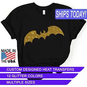 May include: Black t-shirt with a gold glitter bat design. The shirt says "SHIPS TODAY!" in a blue banner at the top and "MADE IN THE USA" in a red, white, and blue banner at the bottom. The shirt also says "CUSTOM DESIGNED HEAT TRANSFERS", "12 GLITTER COLORS", and "MULTIPLE SIZES" in blue banners.