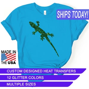 May include: A turquoise blue t-shirt with a green glitter gecko design. The shirt has the text "SHIPS TODAY!" in a purple box at the top right corner. The text "MADE IN THE USA" is in a red, white, and blue box at the bottom left corner. The text "CUSTOM DESIGNED HEAT TRANSFERS" is in a purple box at the bottom center. The text "12 GLITTER COLORS" is in a purple box below the previous text. The text "MULTIPLE SIZES" is in a purple box below the previous text.