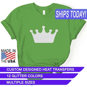 May include: Green t-shirt with a white glitter crown design. The shirt says "SHIPS TODAY!" in blue text. The shirt also says "MADE IN THE USA" with a small American flag. The shirt also says "CUSTOM DESIGNED HEAT TRANSFERS", "12 GLITTER COLORS", and "MULTIPLE SIZES" in purple text.