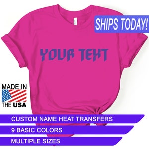 May include: A pink t-shirt with the text "YOUR TEXT" in a purple, bold, blocky font. The shirt is made in the USA and is available in multiple sizes and colors.