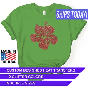 May include: Green t-shirt with a red glitter hibiscus flower design. The text "SHIPS TODAY!" is in a blue box at the top of the image. The text "MADE IN THE USA" is in a red, white, and blue box at the bottom left of the image. The text "CUSTOM DESIGNED HEAT TRANSFERS" is in a purple box at the bottom of the image. The text "12 GLITTER COLORS" is in a purple box below the previous text. The text "MULTIPLE SIZES" is in a purple box below the previous text.