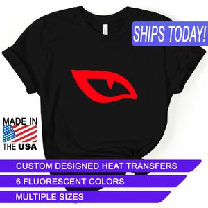 May include: Black t-shirt with a red graphic of a stylized eye with fangs. The shirt has the text "MADE IN THE USA" and "SHIPS TODAY!". Additional text includes "CUSTOM DESIGNED HEAT TRANSFERS", "6 FLUORESCENT COLORS", and "MULTIPLE SIZES".