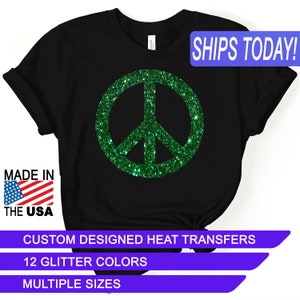 May include: Black t-shirt with a green glitter peace sign. The text "SHIPS TODAY!" is in a blue box at the top of the image. The text "MADE IN THE USA" is in a red, white, and blue box at the bottom left of the image. The text "CUSTOM DESIGNED HEAT TRANSFERS" is in a white box at the bottom of the image. The text "12 GLITTER COLORS" is in a white box below the previous text. The text "MULTIPLE SIZES" is in a white box below the previous text.