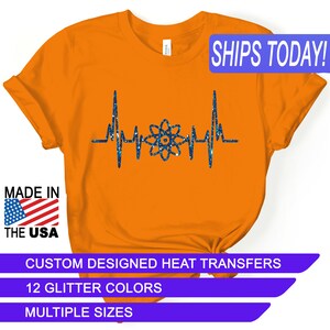 May include: Orange t-shirt with a blue glitter design of an atom with a heartbeat around it. The text "SHIPS TODAY!" is in a blue box at the top of the image. The text "MADE IN THE USA" is in a blue box at the bottom left of the image. The text "CUSTOM DESIGNED HEAT TRANSFERS" is in a blue box at the bottom of the image. The text "12 GLITTER COLORS" is in a blue box below the previous text. The text "MULTIPLE SIZES" is in a blue box below the previous text.