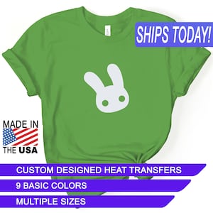 May include: Green t-shirt with a white bunny rabbit graphic. The shirt has the text "SHIPS TODAY!" in a blue box at the top right corner. The text "MADE IN THE USA" is in a red, white, and blue box at the bottom left corner. The text "CUSTOM DESIGNED HEAT TRANSFERS" is in a purple box at the bottom center. The text "9 BASIC COLORS" is in a purple box below the previous text. The text "MULTIPLE SIZES" is in a purple box below the previous text.