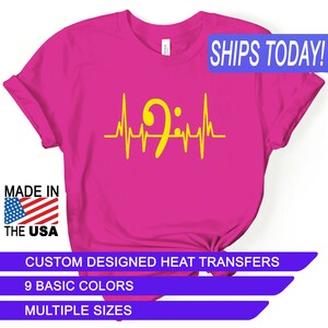May include: A pink t-shirt with a yellow bass clef and heartbeat graphic. The text "SHIPS TODAY!" is in a blue box at the top of the image. The text "MADE IN THE USA" is in a red, white, and blue box at the bottom left of the image. The text "CUSTOM DESIGNED HEAT TRANSFERS" is in a purple box at the bottom of the image. The text "9 BASIC COLORS" is in a purple box below the previous text. The text "MULTIPLE SIZES" is in a purple box below the previous text.