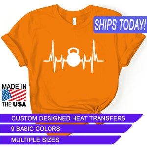 May include: Orange t-shirt with a white graphic of a kettlebell inside a heartbeat. The text "SHIPS TODAY!" is in a blue box at the top of the image. The text "MADE IN THE USA" is in a red, white, and blue box at the bottom left of the image. The text "CUSTOM DESIGNED HEAT TRANSFERS" is in a purple box at the bottom of the image. The text "9 BASIC COLORS" is in a purple box below the previous text. The text "MULTIPLE SIZES" is in a purple box below the previous text.