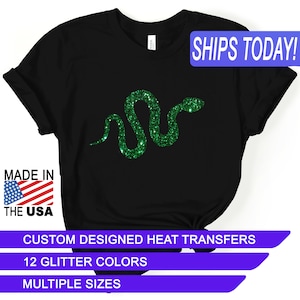 May include: Black t-shirt with a green glitter snake design. The text "SHIPS TODAY!" is in a blue box at the top of the image. The text "MADE IN THE USA" is in a red, white, and blue box at the bottom left of the image. The text "CUSTOM DESIGNED HEAT TRANSFERS" is in a blue box at the bottom of the image. The text "12 GLITTER COLORS" is in a blue box below the previous text. The text "MULTIPLE SIZES" is in a blue box below the previous text.