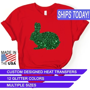 May include: Red t-shirt with a green glitter bunny silhouette. The text "SHIPS TODAY!" is in a blue box at the top of the image. The text "MADE IN THE USA" is in a blue box at the bottom left of the image. The text "CUSTOM DESIGNED HEAT TRANSFERS" is in a blue box at the bottom center of the image. The text "12 GLITTER COLORS" is in a blue box below the previous text. The text "MULTIPLE SIZES" is in a blue box below the previous text.