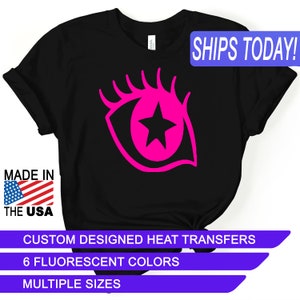 May include: Black t-shirt with a pink eye design featuring a black star in the center. The text "SHIPS TODAY!" is in a blue box at the top of the image. The text "MADE IN THE USA" is in a red, white, and blue box at the bottom left of the image. The text "CUSTOM DESIGNED HEAT TRANSFERS" is in a purple box at the bottom of the image. The text "6 FLUORESCENT COLORS" is in a purple box below the previous text. The text "MULTIPLE SIZES" is in a purple box below the previous text.