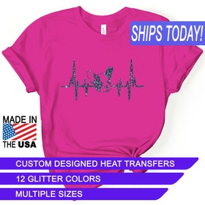 May include: Pink t-shirt with a glitter design of a dog's head with a heartbeat line. The text "SHIPS TODAY!" is in a blue box at the top of the image. The text "MADE IN THE USA" is in a red, white, and blue box at the bottom left of the image. The text "CUSTOM DESIGNED HEAT TRANSFERS" is in a white box at the bottom of the image. The text "12 GLITTER COLORS" is in a white box below the previous text. The text "MULTIPLE SIZES" is in a blue box below the previous text.