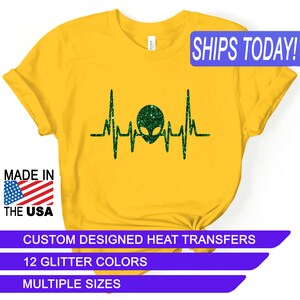 May include: Yellow t-shirt with a green glitter alien head inside a heartbeat graphic. The text "SHIPS TODAY!" is in a blue box at the top of the image. The text "MADE IN THE USA" is in a red, white, and blue box at the bottom left of the image. The text "CUSTOM DESIGNED HEAT TRANSFERS" is in a purple box at the bottom of the image. The text "12 GLITTER COLORS" is in a purple box below the previous text. The text "MULTIPLE SIZES" is in a purple box below the previous text.