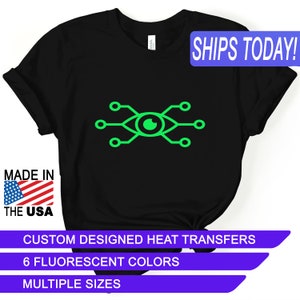 May include: Black t-shirt featuring a neon green eye design with circuit-like elements. The shirt includes the text "MADE IN THE USA". Additional text reads "CUSTOM DESIGNED HEAT TRANSFERS", "6 FLUORESCENT COLORS", and "MULTIPLE SIZES".