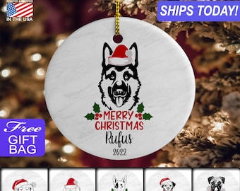 May include: White ceramic Christmas ornament with a black and white German Shepherd dog wearing a red Santa hat. The ornament has the text "Merry Christmas Rufus 2022" on it.