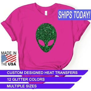 May include: Pink t-shirt with a green glitter alien head design. The text "SHIPS TODAY!" is in a blue box at the top of the image. The text "MADE IN THE USA" is in a red, white, and blue box at the bottom left of the image. The text "CUSTOM DESIGNED HEAT TRANSFERS" is in a blue box at the bottom of the image. The text "12 GLITTER COLORS" is in a blue box below the previous text. The text "MULTIPLE SIZES" is in a blue box below the previous text.