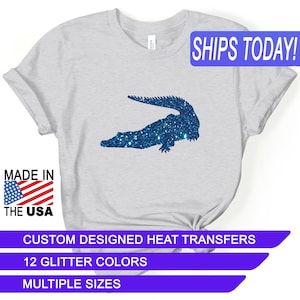 May include: A gray t-shirt with a blue glitter alligator design. The shirt says "SHIPS TODAY!" in a blue banner at the top. The shirt also says "MADE IN THE USA" with a small American flag. The shirt also says "CUSTOM DESIGNED HEAT TRANSFERS", "12 GLITTER COLORS", and "MULTIPLE SIZES" in blue banners at the bottom.