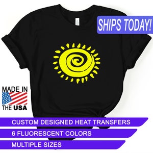 May include: Black t-shirt with a yellow sun graphic. The sun has a spiral design in the center. The text "SHIPS TODAY!" is in the upper right corner. The text "MADE IN THE USA" is in the lower left corner. The text "CUSTOM DESIGNED HEAT TRANSFERS" is in the lower center. The text "6 FLUORESCENT COLORS" is in the lower center. The text "MULTIPLE SIZES" is in the lower center.