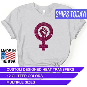 May include: A gray t-shirt with a pink glitter iron-on design of a raised fist inside a female symbol. The text "SHIPS TODAY!" is in a blue box at the top of the image. The text "MADE IN THE USA" is in a red, white, and blue box at the bottom left of the image. The text "CUSTOM DESIGNED HEAT TRANSFERS" is in a purple box at the bottom of the image. The text "12 GLITTER COLORS" is in a purple box below the previous text. The text "MULTIPLE SIZES" is in a purple box below the previous text.