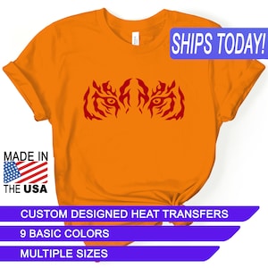 May include: Orange t-shirt with a red tiger eye design. The text "SHIPS TODAY!" is in a blue box at the top of the image. The text "MADE IN THE USA" is in a blue box at the bottom left of the image. The text "CUSTOM DESIGNED HEAT TRANSFERS" is in a purple box at the bottom of the image. The text "9 BASIC COLORS" is in a purple box below the previous text. The text "MULTIPLE SIZES" is in a purple box below the previous text.