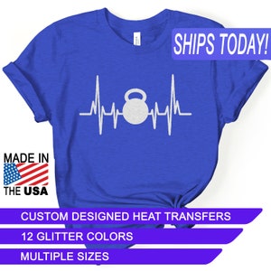 May include: Royal blue t-shirt featuring a white graphic of a kettlebell within a heartbeat line. The text "MADE IN THE USA" is visible. Additional text includes "CUSTOM DESIGNED HEAT TRANSFERS", "12 GLITTER COLORS", and "MULTIPLE SIZES".