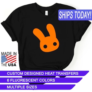 May include: Black t-shirt with an orange bunny rabbit graphic. The text "SHIPS TODAY!" is in a blue box at the top of the image. The text "MADE IN THE USA" is in a red, white, and blue box at the bottom left of the image. The text "CUSTOM DESIGNED HEAT TRANSFERS", "6 FLUORESCENT COLORS", and "MULTIPLE SIZES" are in purple boxes at the bottom of the image.