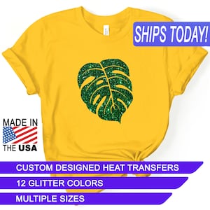 May include: A yellow t-shirt with a green glitter monstera leaf design. The shirt has the text "SHIPS TODAY!" in a blue box at the top right corner. The text "MADE IN THE USA" is in a red, white, and blue box at the bottom left corner. The text "CUSTOM DESIGNED HEAT TRANSFERS" is in a purple box at the bottom center. The text "12 GLITTER COLORS" is in a purple box below the previous text. The text "MULTIPLE SIZES" is in a purple box below the previous text.