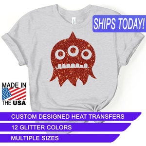 May include: A white t-shirt with a red glitter three-eyed monster design. The text "SHIPS TODAY!" is in a blue box at the top of the image. The text "MADE IN THE USA" is in a red, white, and blue box at the bottom left of the image. The text "CUSTOM DESIGNED HEAT TRANSFERS" is in a blue box at the bottom of the image. The text "12 GLITTER COLORS" is in a blue box below the previous text. The text "MULTIPLE SIZES" is in a blue box below the previous text.