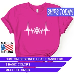 May include: Pink t-shirt with a white atom symbol inside a heartbeat graphic. The text "SHIPS TODAY!" is in a blue box at the top of the image. The text "MADE IN THE USA" is in a red, white, and blue box at the bottom left of the image. The text "CUSTOM DESIGNED HEAT TRANSFERS" is in a blue box at the bottom of the image. The text "9 BASIC COLORS" is in a blue box below the previous text. The text "MULTIPLE SIZES" is in a blue box below the previous text.