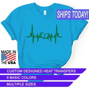 May include: A turquoise blue t-shirt with a green heart beat graphic featuring an open book in the center. The text "SHIPS TODAY!" is in a purple box at the top of the image. The text "MADE IN THE USA" is in a red, white, and blue box at the bottom left of the image. The text "CUSTOM DESIGNED HEAT TRANSFERS" is in a purple box at the bottom of the image. The text "9 BASIC COLORS" is in a purple box below the previous text. The text "MULTIPLE SIZES" is in a purple box below the previous text.