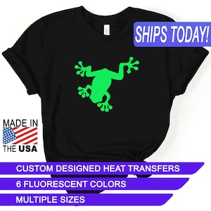 May include: Black t-shirt with a green frog silhouette. The text "SHIPS TODAY!" is in a blue box at the top of the image. The text "MADE IN THE USA" is in a red, white, and blue box at the bottom left of the image. The text "CUSTOM DESIGNED HEAT TRANSFERS" is in a purple box at the bottom of the image. The text "6 FLUORESCENT COLORS" is in a purple box below the previous text. The text "MULTIPLE SIZES" is in a purple box below the previous text.
