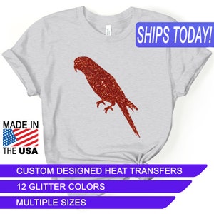 May include: A gray t-shirt with a red glitter parrot silhouette. The shirt has the text "SHIPS TODAY!" in red letters on the top right corner. The text "MADE IN THE USA" is on the bottom left corner. The text "CUSTOM DESIGNED HEAT TRANSFERS" is on the bottom center of the shirt. The text "12 GLITTER COLORS" is on the bottom center of the shirt. The text "MULTIPLE SIZES" is on the bottom center of the shirt.
