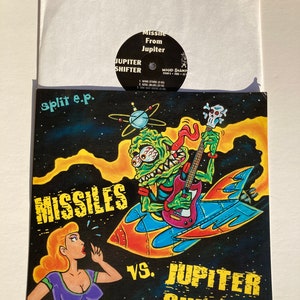 May include: A split 7-inch vinyl record with a black and white illustration of a green monster playing a guitar on a rocket ship. The text "MISSILES VS. JUPITER SHIFTER" is printed in yellow on a black background. The record is titled "split e.p."