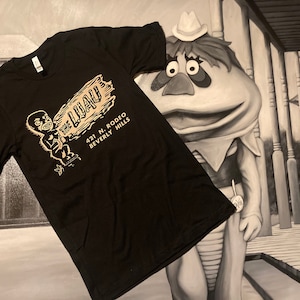 Puede incluir: Camiseta negra con un gráfico blanco de un personaje de dibujos animados sosteniendo un cartel que dice "The Luau" con la dirección "421 N. Rodeo Beverly Hills".