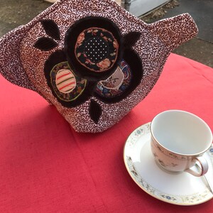 Könnte beinhalten: Ein brauner und weißer Teekannenwärmer mit Blumenmuster. Der Wärmer hat drei große Kreise mit verschiedenen Mustern, darunter Streifen, Punkte und Paisley. Der Wärmer steht auf einer roten Tischdecke neben einer weißen Teetasse und Untertasse.