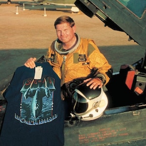 Puede incluir: Un hombre con un traje de vuelo amarillo se sienta en la cabina de un avión SR-71 Blackbird negro. Sostiene una camiseta azul con una imagen en blanco y negro del SR-71 Blackbird. El texto "SR-71 Blackbird" está impreso en la camiseta.