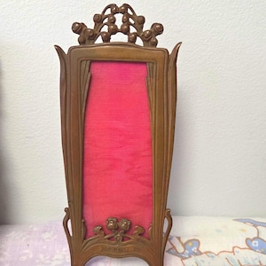 May include: A tall, vertical bronze-toned metal picture frame featuring an ornate, flowing design with floral motifs at the top and bottom. The frame surrounds a vibrant, textured hot pink fabric insert. The word Mermé is embossed at the base.