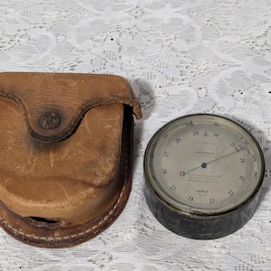 Puede incluir: Una funda de cuero vintage con un barómetro aneroide redondo de cara plateada con una aguja negra y el texto "The Spring Compensated Aneroid Barometer Made in England" en la cara.