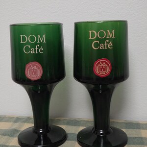 Puede incluir: Dos copas verdes con la inscripción "DOM Café" en letras doradas. Cada copa tiene un emblema rojo y dorado. Las copas están sobre una superficie a cuadros.