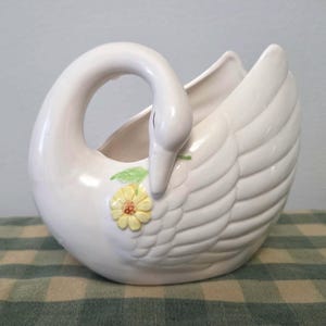Peut inclure: Un pot de fleurs en forme de cygne en céramique blanche, avec un cou incurvé et des ailes détaillées. Une fleur jaune avec des feuilles vertes est sur le devant. Le pot est sur une nappe à carreaux verts et blancs.