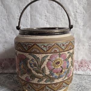 Peut inclure: Un pot en céramique avec un motif floral rose, bleu et jaune, agrémenté de feuilles vertes. Le pot a un bord et une poignée en métal argenté et une base dorée. Le motif floral est sur fond crème.