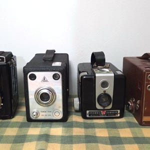 May include: Four vintage box cameras of varying designs. The cameras are black and brown, with details in silver and tan. One camera has the text "Lubitel 2" on top. The cameras are arranged on a checkered surface.