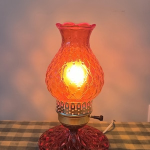 May include: A vintage red glass lamp with a textured shade and base. The lamp has a ruffled top and a brass-coloured base. The lamp is lit, emitting a warm orange glow. The lamp is on a checkered tablecloth.