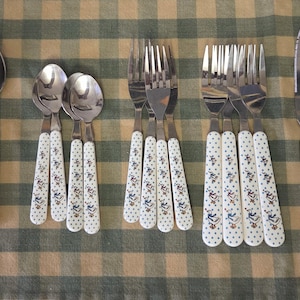 Könnte beinhalten: Ein Set aus Vintage-Besteck mit weißen Griffen, die mit blauen und gelben Blumen- und Vogelmustern verziert sind. Das Set umfasst Löffel, Gabeln und Messer, die auf einer karierten Tischdecke angeordnet sind.